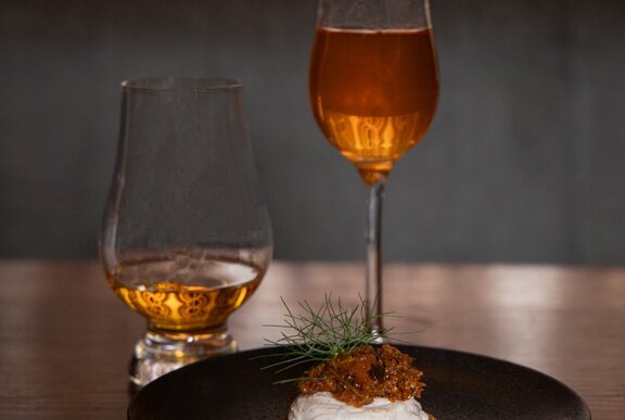A whisky cocktail in a tall champagne glass alongside a nip of whisky in a stemless cognac glass, with a cracker with toppings on a black plate.