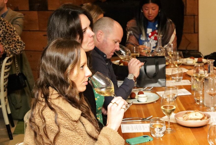 Diners sampling wine at a long wooden table set with glasses and cutlery.