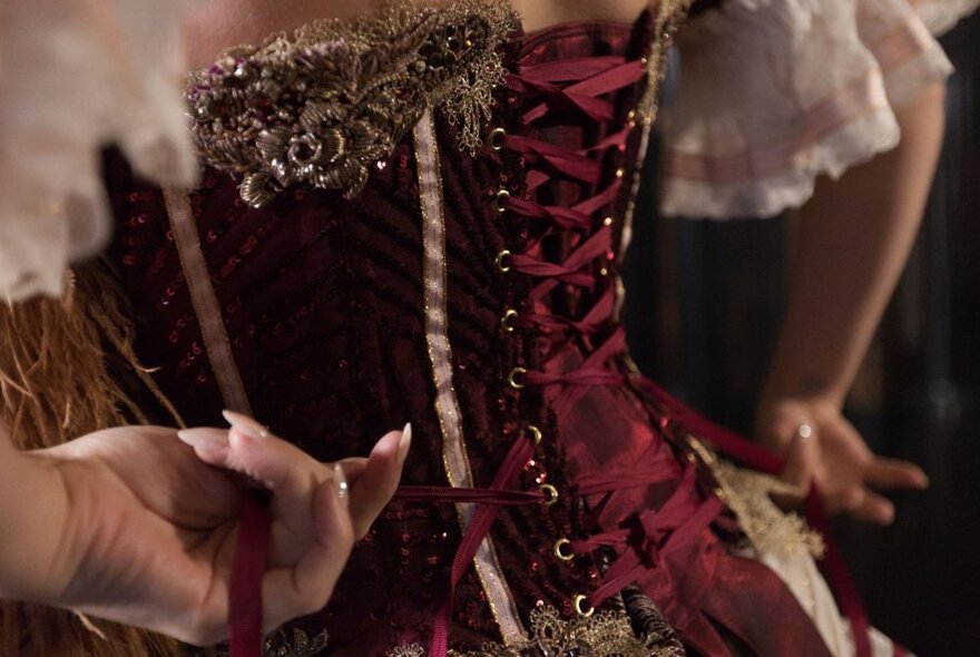 Close up of hands tightening the red laces of a corset being worn by a burlesque dancer.