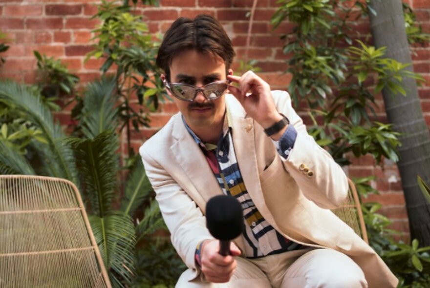 Comedian Sweeney Preston wearing a suit and loud shirt, looking over the top of a pair of sunglasses as he holds a microphone.