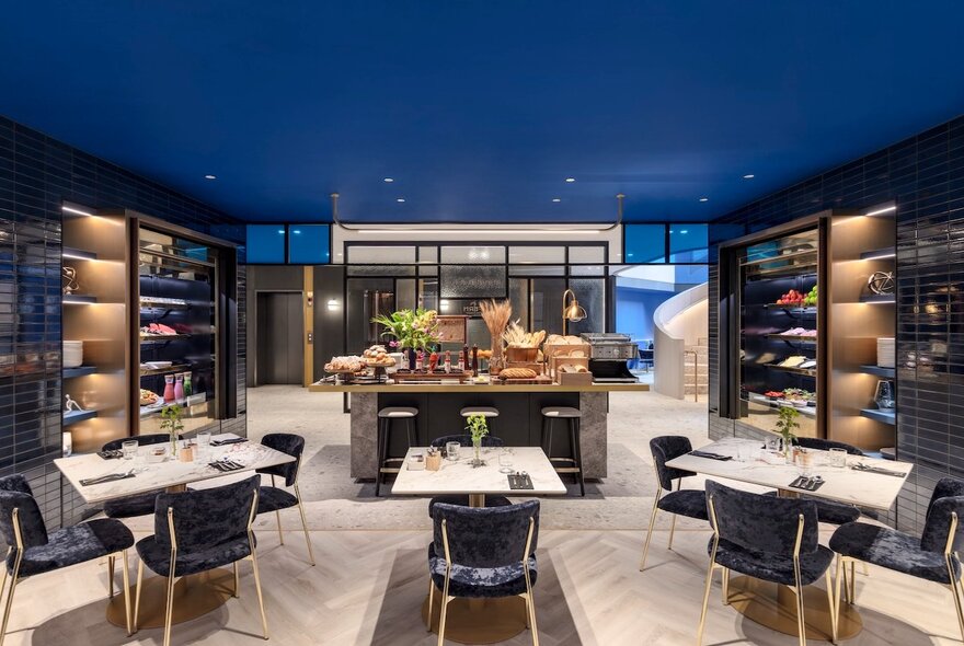Dining area of a hotel, with a deep blue ceiling, pale wooden floor, and a glimpse of the lobby in the background.