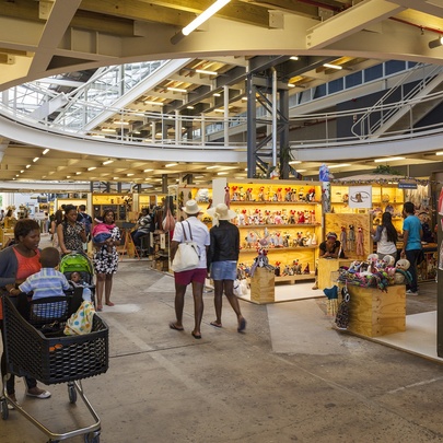 Interior view of a bustling indoor craft market with a large circular ceiling opening, timber structural beams, and shoppers browsing various stalls.