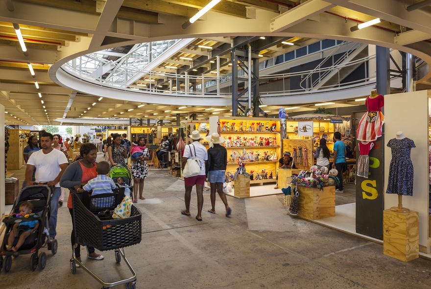 Interior view of a bustling indoor craft market with a large circular ceiling opening, timber structural beams, and shoppers browsing various stalls.