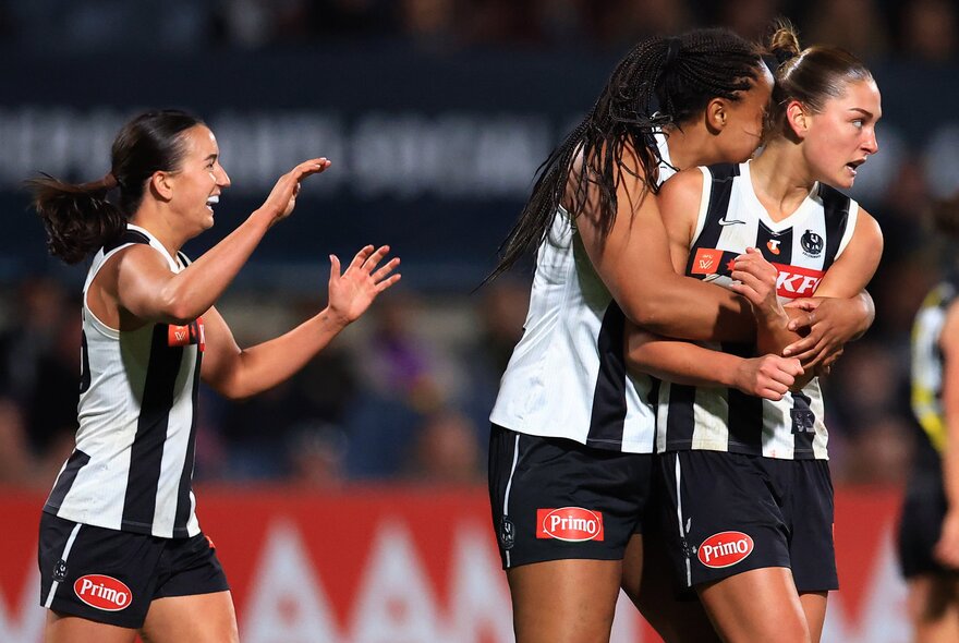 Collingwood AFLW players embrace in celebration on the football field, another smiling with her arms raised. 