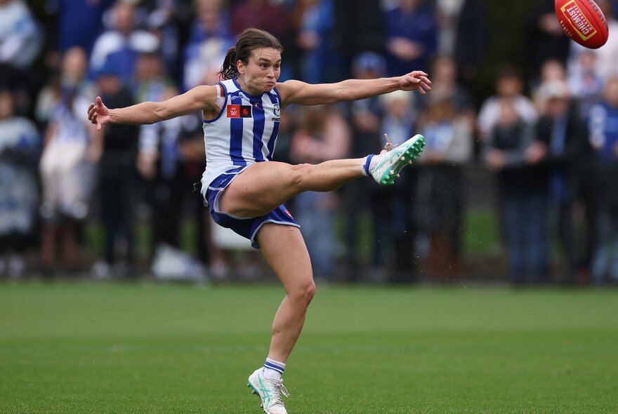 A North Melbourne AFLW player kicking the ball on the football field, her leg high in the air.