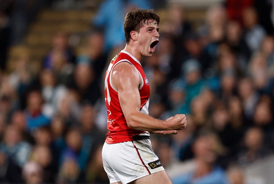 A St Kilda AFL football player with his fists clenched and pumped during a match, a blurred stadium crowd in the background.