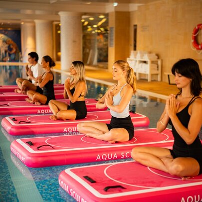 Five women sitting cross-legged on red blow-up pilates mats, floating on the surface of a hotel pool.