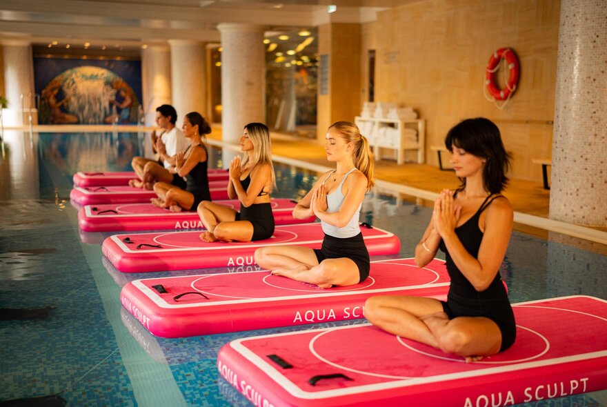 Five women sitting cross-legged on red blow-up pilates mats, floating on the surface of a hotel pool.