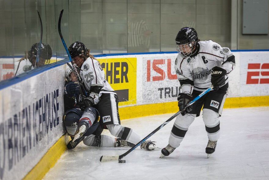 An ice hockey player crashes into another on the side of an indoor ice hockey field, while another player takes control of the puck. 