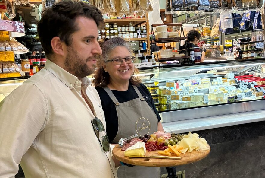 A man holding a wooden charcuterie board with a selection of cheese and meats, a woman in an apron standing next to him; a deli counter in the background.