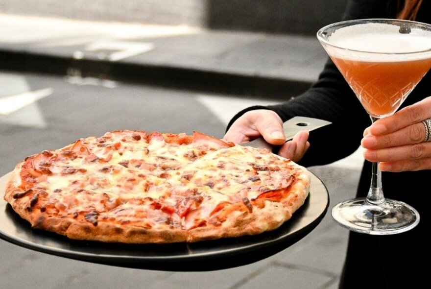 Hands of a waiter visible holding a tray of pizza and a cocktail. 