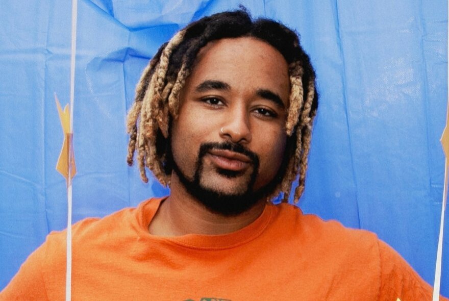 A head-and-shoulders portrait of a man with blonde-tipped dreadlocks, a thin moustache, and a goatee, wearing an orange t-shirt and looking directly forward with a calm expression against a bright blue draped backdrop.