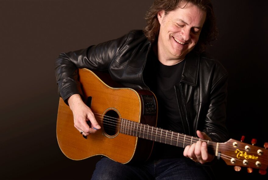 Musician Mark Hughes playing guitar and smiling, looking down at the fretboard.