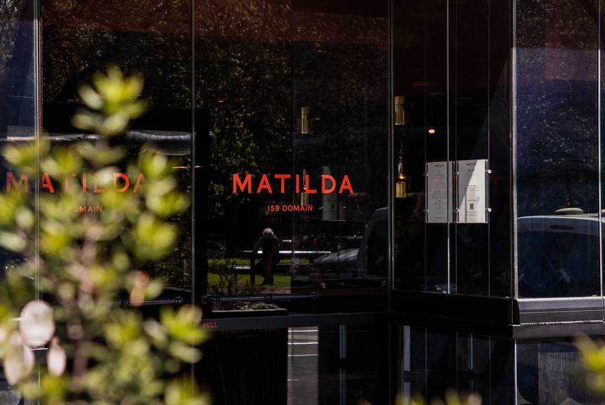 The exterior of the restaurant Matilda, with the red name on a glass door, an out of focus plant in the foreground. 