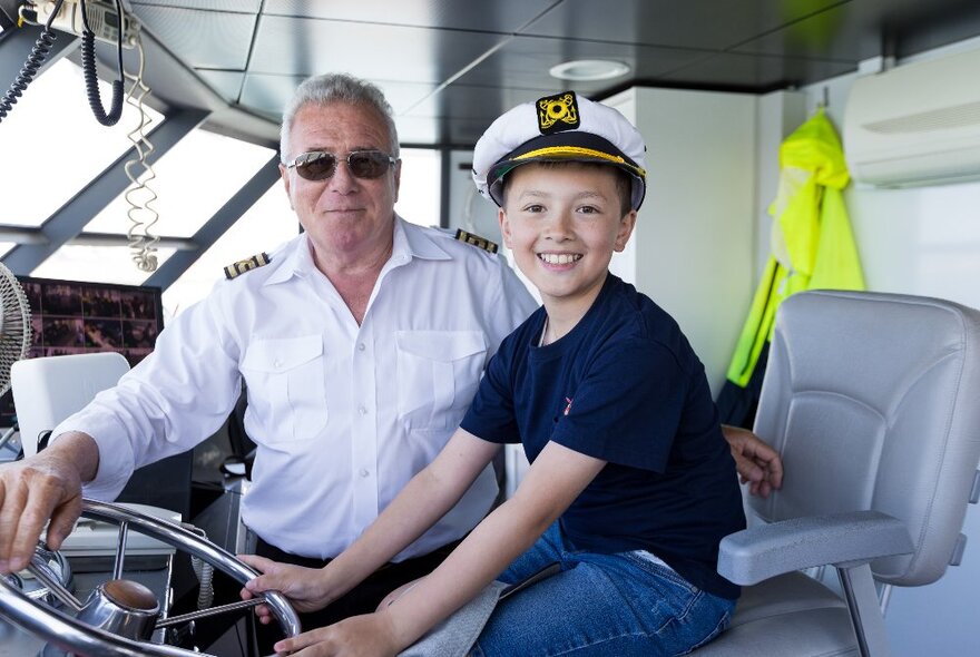 A boy with a captain's hat on having a go at steering a ship, the actual captain next to him. 