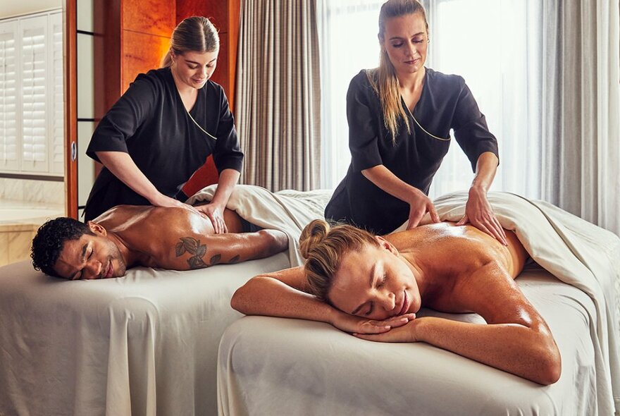 Therapists treating a couple lying on massage tables under blankets.