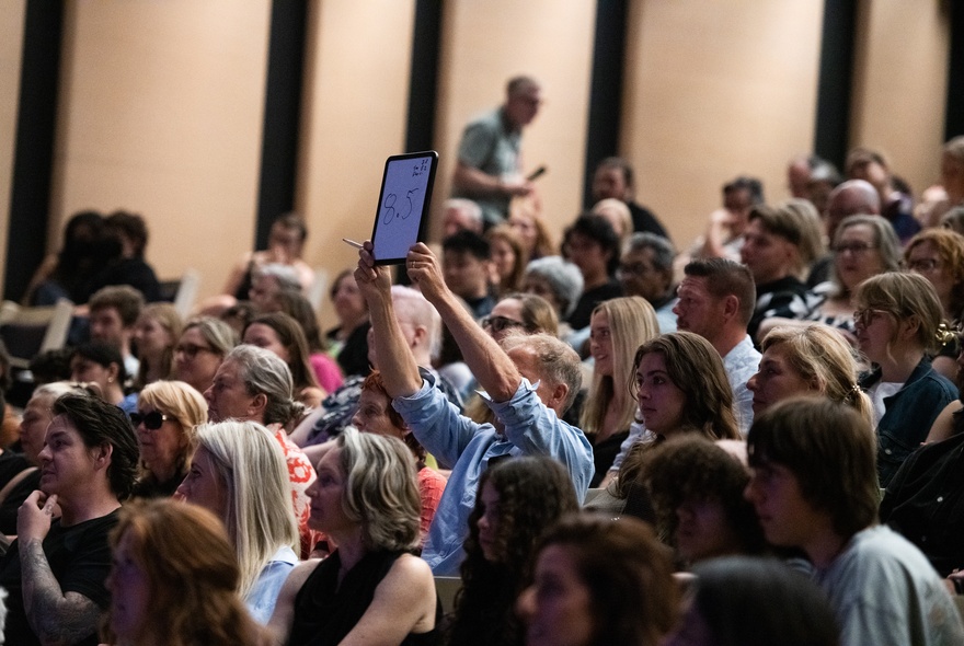 Seated audience in a venue, one person holding up an iPad..