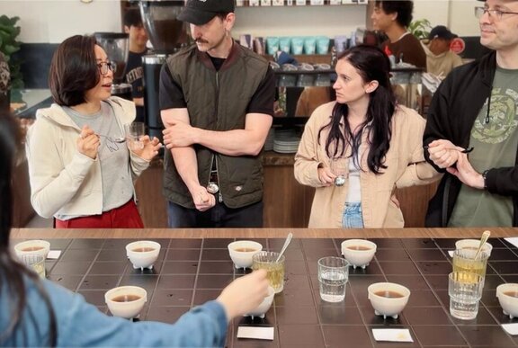 People standing around a table that has several cups of coffee on it, glasses of water and teaspoons, inside a coffee roastery, listening to an expert talk about coffee.