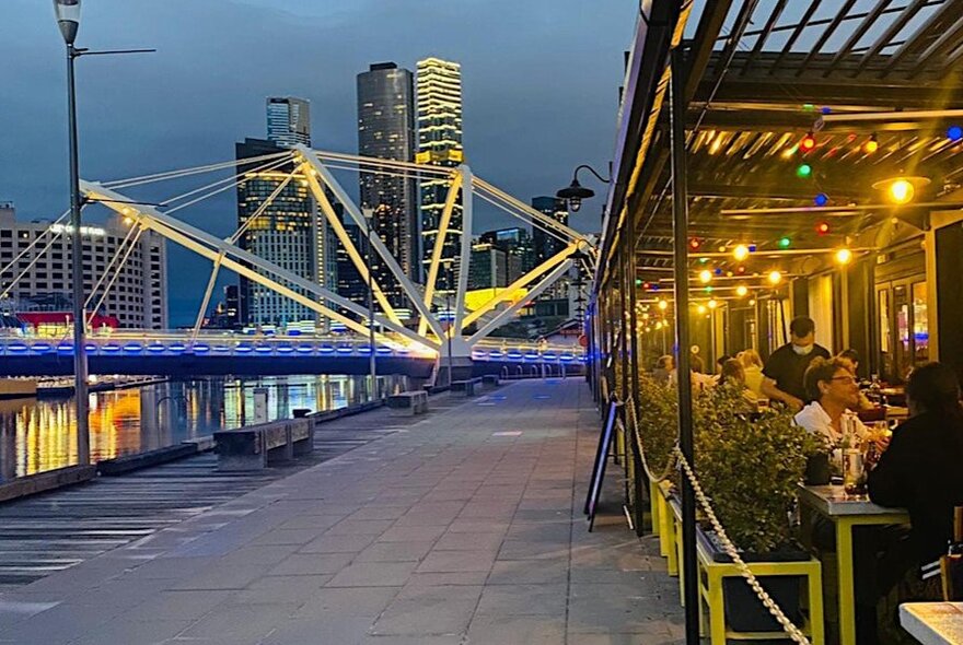 Riverside dining with people seated under an annex and city buildings visible in the distance. 