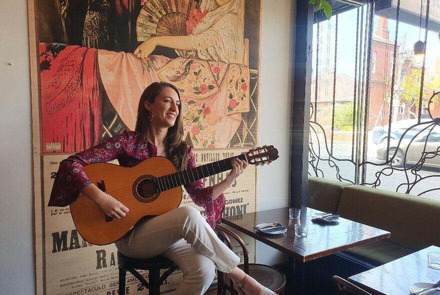 Laura de Elvira playing acoustic guitar indoors at a small restaurant table by a window.