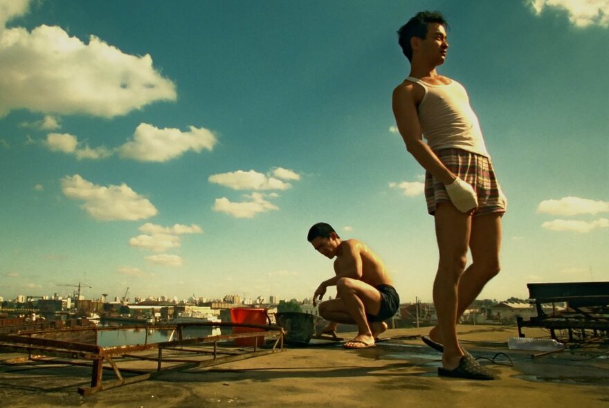 A still from a Hong Kong film showing two men, one crouching and one standing and leaning back, with a blue sky and clouds behind them. 