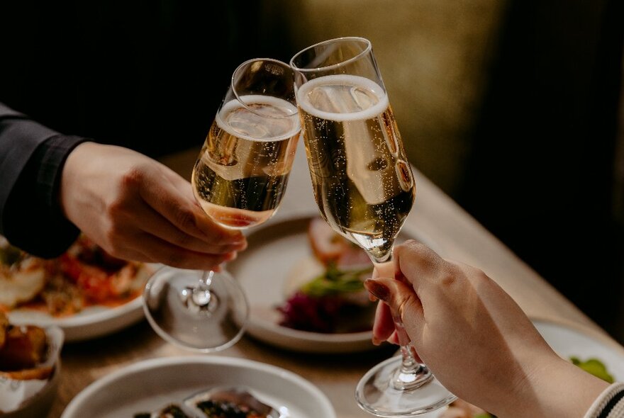 Two hands holding champagne flute glasses,  bringing them together across a table in a celebratory clinking motion.