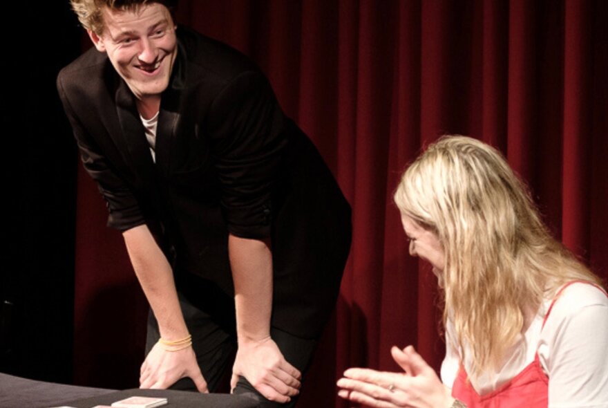 Smiling magician with his hands on his knees, next to a blonde woman applauding.