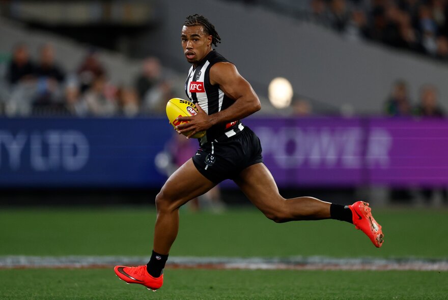 A Collingwood AFL player running on the field, a yellow football he holds in his hands, with blurred fans in the stadium in the background.