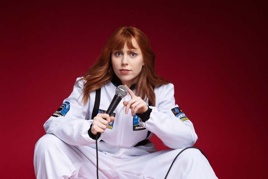 Performer Ally Morgan in a white space suit, holding a microphone, against a deep red background. 
