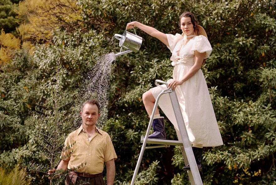 A woman on a stepladder in a garden pours water from a watering can over a man standing below.