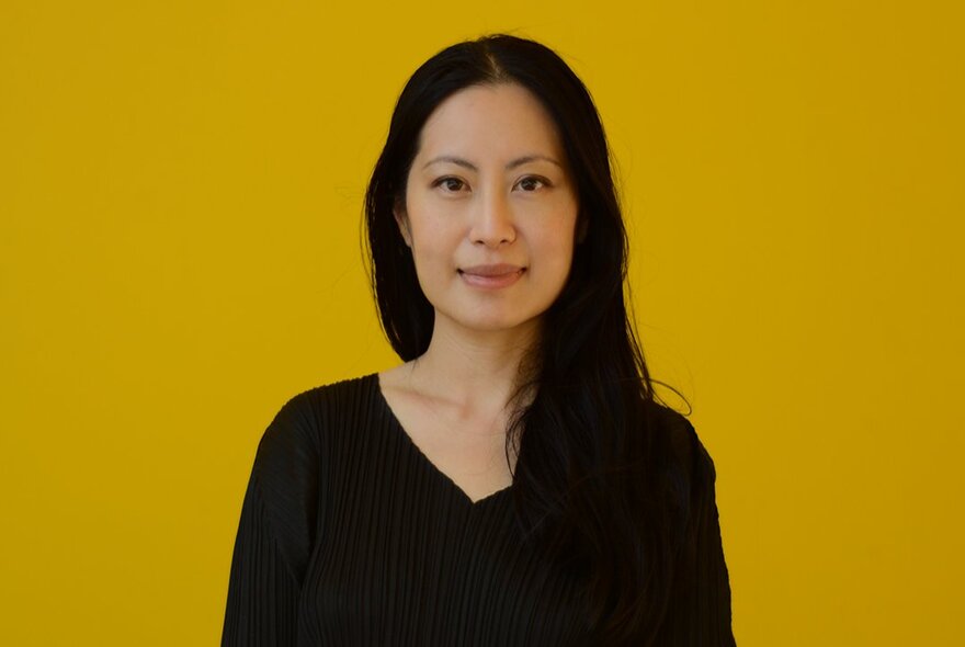 Monica Lim, a woman with long dark hair, smiling slightly, wearing a v-neck top, posed against a dark yellow background.