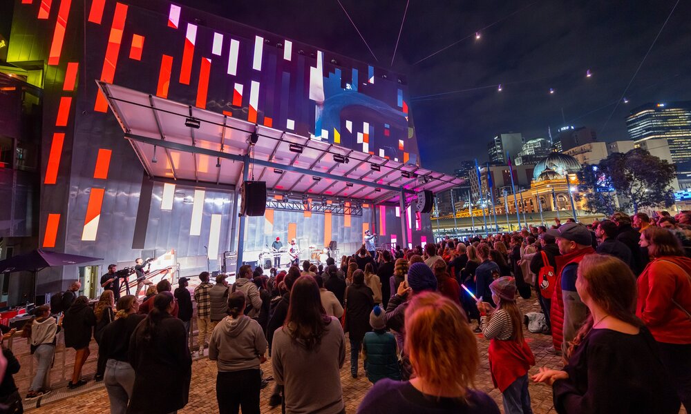 A crowd of people are watching a band play on a stage at Federation Square at night.