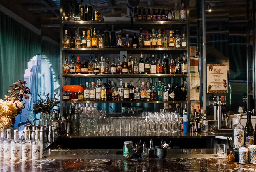 A dimly lit bar counter featuring a multi-tiered display of liquor bottles on shelves in the background.