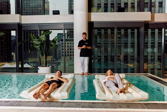 Two people meditating on pool floats with an instructor on the edge of the pool between them. 