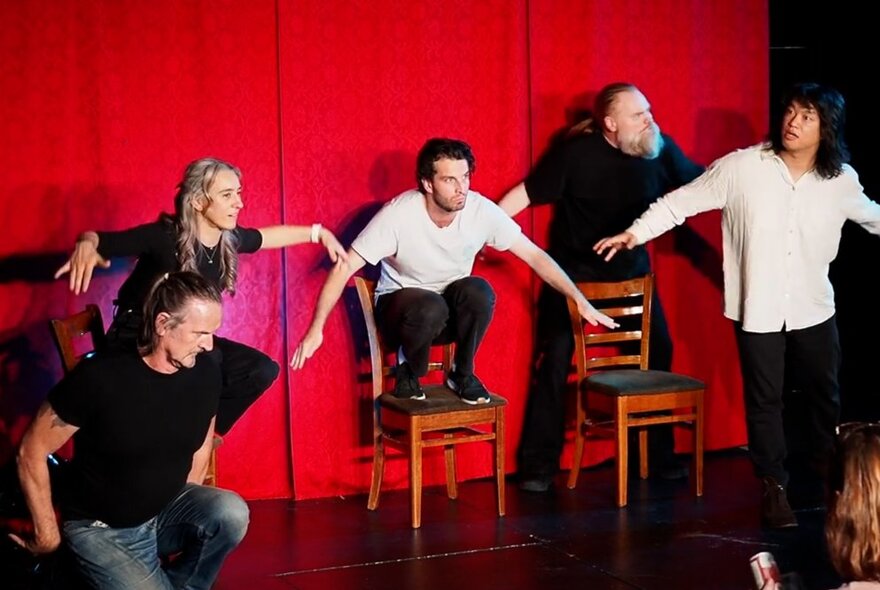 Five performers in close proximity improvising on a stage in front of a red curtain, using chairs as props.