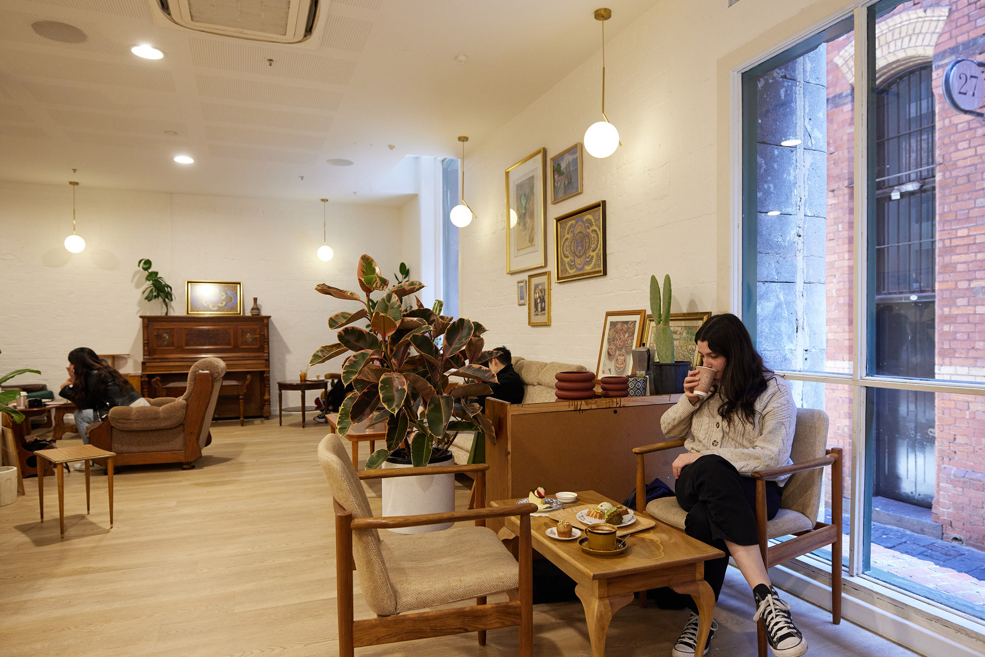 A person is sitting in a cafe with retro furniture and plants. They are drinking coffee there is a table with cakes and desserts on it