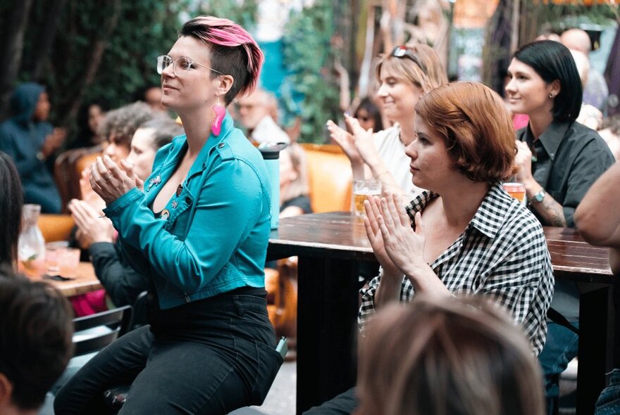 A small crowd of people seated at tables and on benches, clapping.