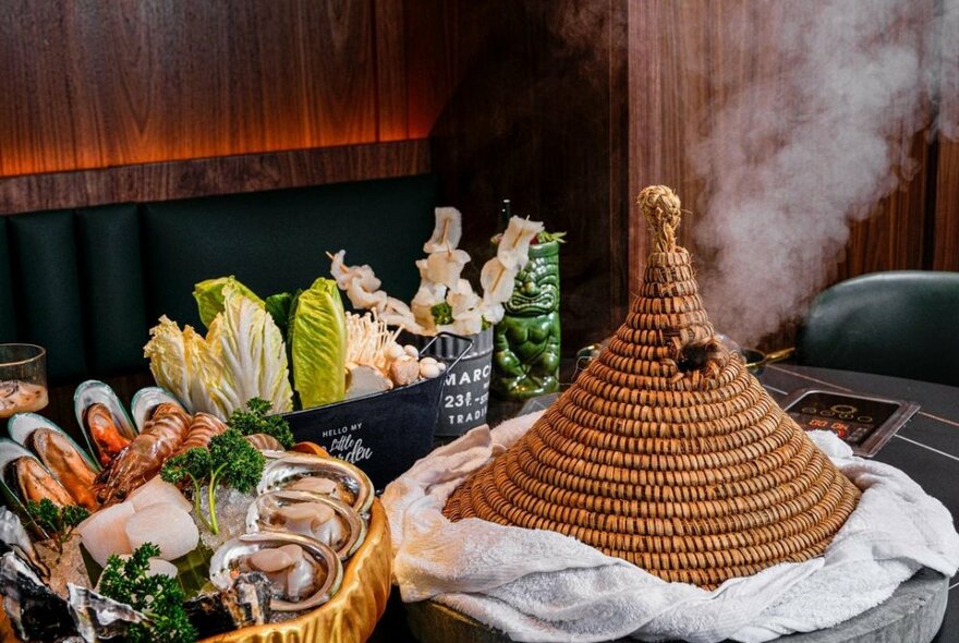 Conical woven pot lid covering a stone cooking pot at a table next to baskets filled with seafood and vegetables.