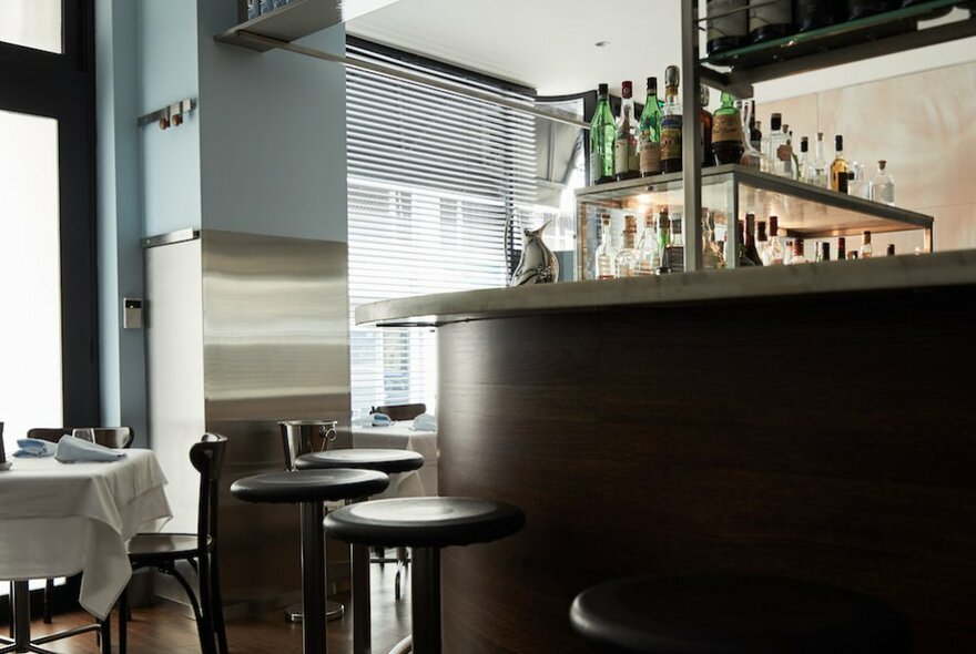 Interior of restaurant showing high counter with wine bottles, bar stools and a small table near the window set for a dinner service.