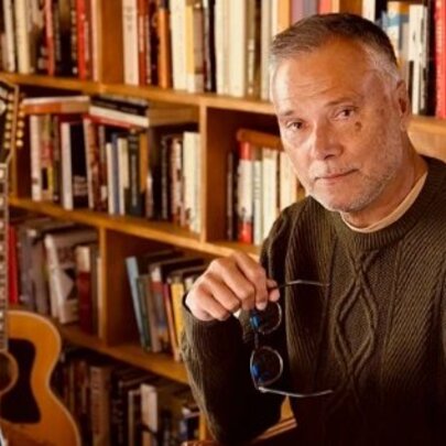 Portrait of TV presenter and author Stan Grant in his home library, sitting in front of his computer with his glasses in his hand.