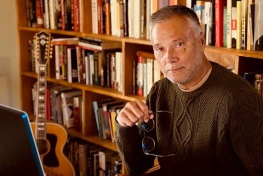 Portrait of TV presenter and author Stan Grant in his home library, sitting in front of his computer with his glasses in his hand.