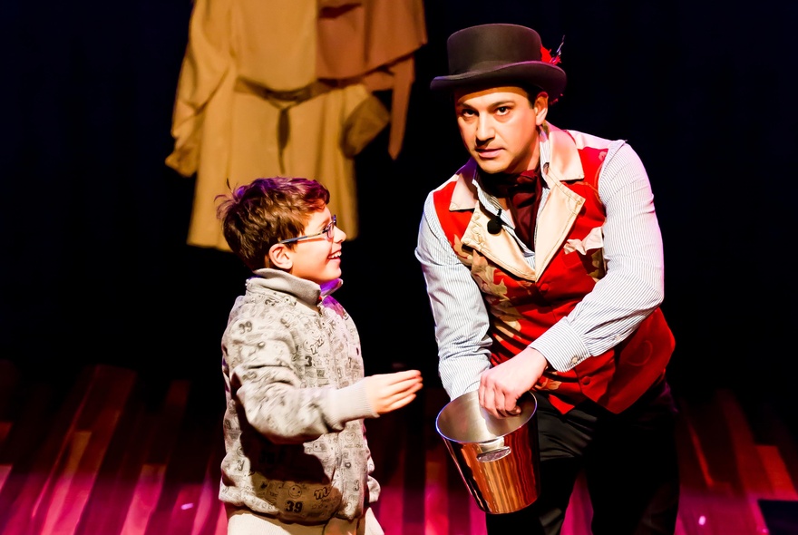 A magician wearing a top hat, on a stage, interacting with a primary school aged child.