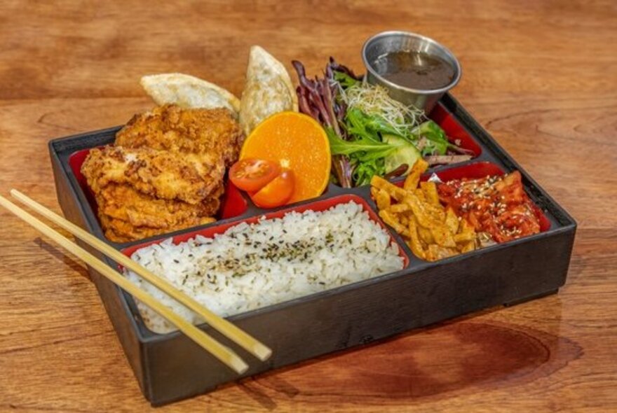 A bento box with Korean chicken, rice, salad and other bites, a set of chopsticks balanced over the lower left corner. 