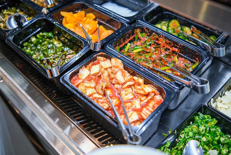 Selection of Korean side dishes in rectangular serving dishes with tongs, on a buffet table.