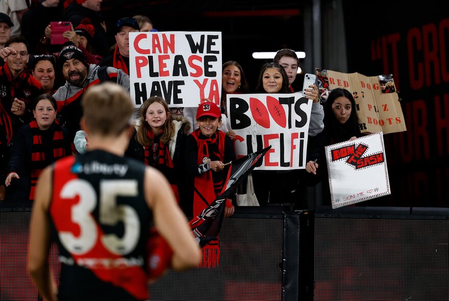 The blurred back of an Essendon player as he walks towards a crowd of supporters with banners. 