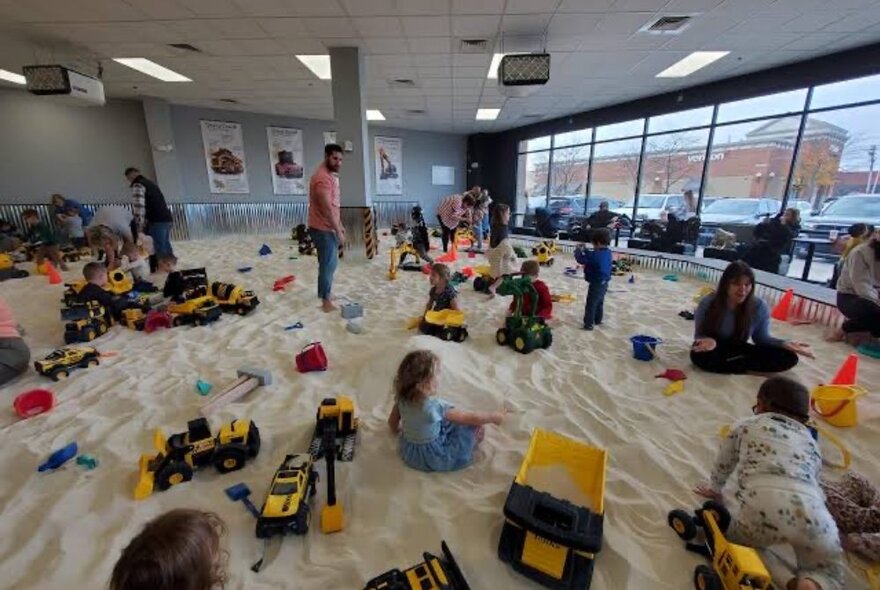 Children in a large indoor sandpit filled with dump-trucks and diggers. 