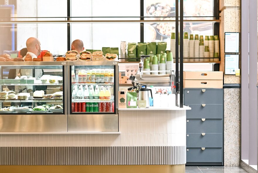Brightly lit cafe counter with bar fridge and rows of takeaway food including sandwiches.