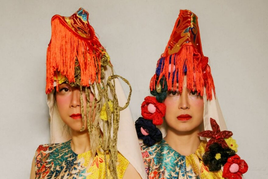 Two women, each wearing brightly coloured tops and elaborate red and orange headdresses, with hanging decorative ornaments and floral motifs, that partly covers their faces.