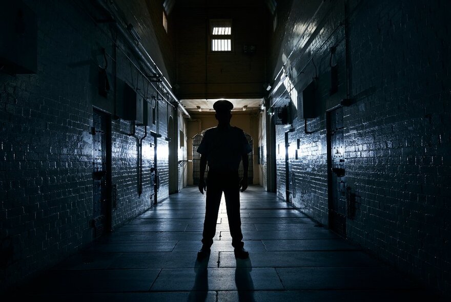 Dark figure standing in a dark corridor lined with closed cell doors in a jail.