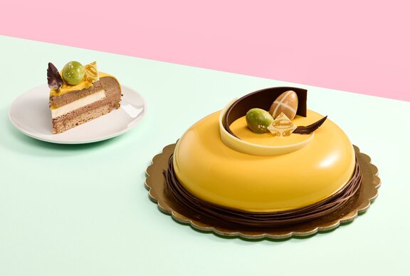 A smooth, yellow frosted, round cake on a dark cake plate, next to a small slice of layered cake on a separate plate. 